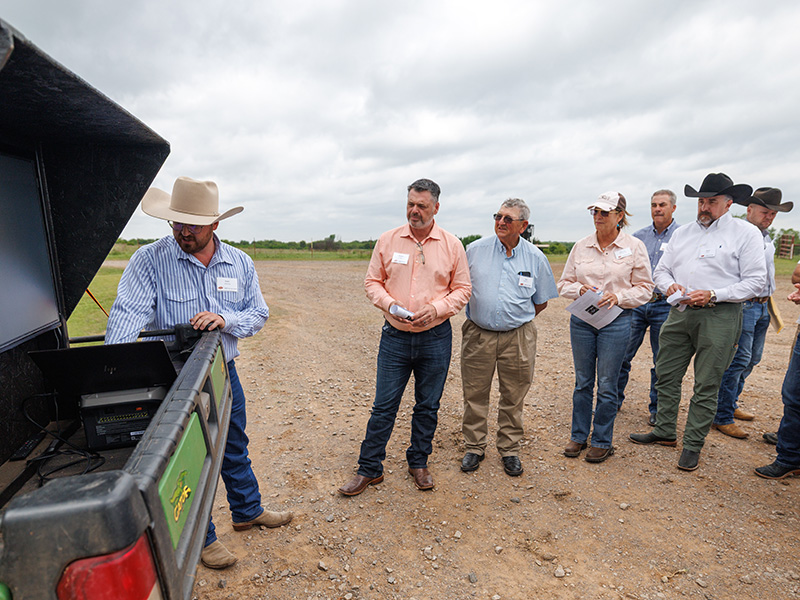 A presenter demonstrates technology from the back of a pickup truck while a group of attendees in ranch attire observe during an outdoor agricultural field event.