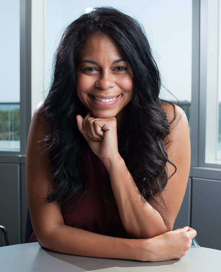 A person sits at a table with one hand resting beneath the chin, looking toward the camera in a professional portrait taken in front of large windows.