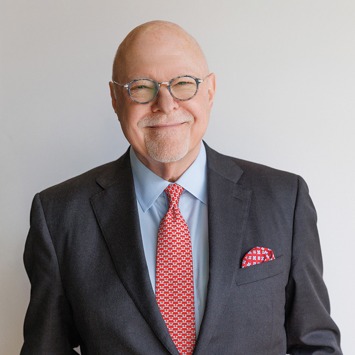 A person wearing a dark suit, light blue shirt and patterned red tie poses for a professional portrait against a plain light-colored background.