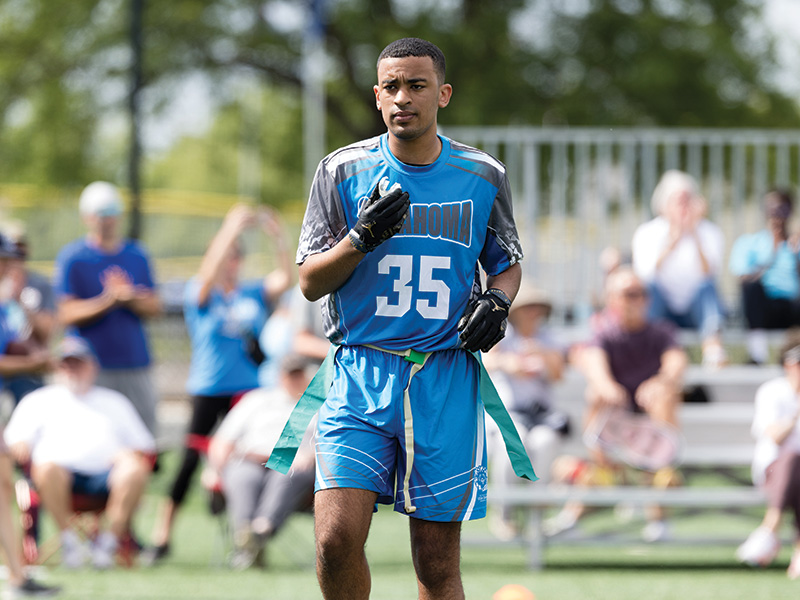 An athlete wearing an Oklahoma flag football uniform stands on the field during competition, with spectators seated in the background and green flag belts visible at the waist.
