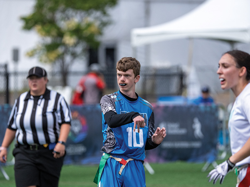 An Oklahoma flag football player wearing jersey number 10 gestures to teammates during a game while a referee and opposing player stand nearby on the field.