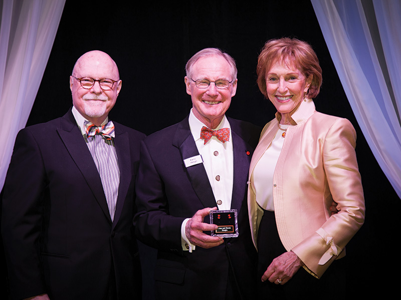 Three people in formal attire pose together at an awards event, with the person in the center holding a presentation box while standing in front of draped stage décor.
