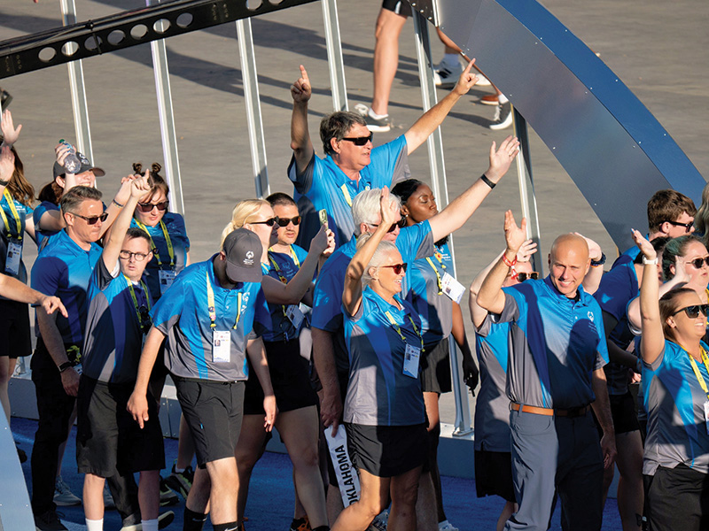 A group of athletes and coaches in matching blue uniforms wave and walk together during an opening ceremony or team parade at a large sporting event.