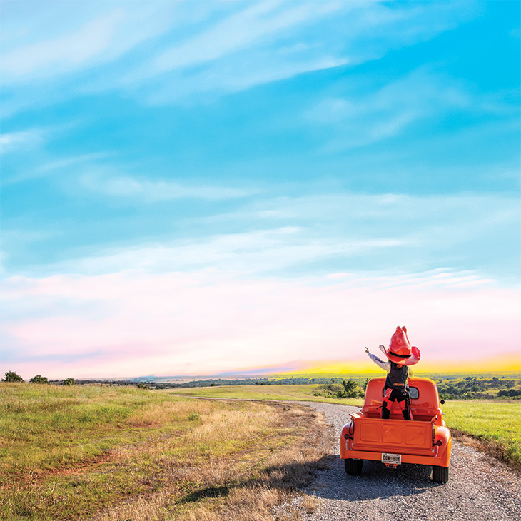 Pistol Pete rides in the back of an orange pickup truck on a winding gravel road through open grasslands, pointing toward the horizon beneath a colorful sunset sky.