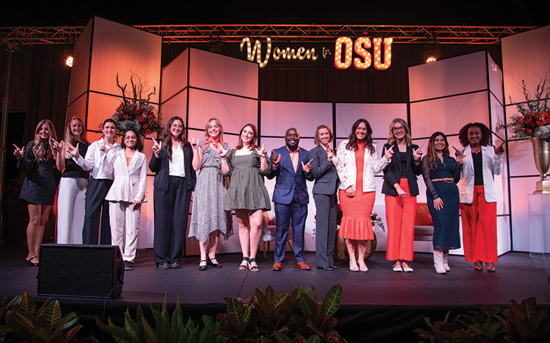 A group of scholarship recipients and event organizers pose together on stage beneath a “Women for OSU” sign during a recognition ceremony.
