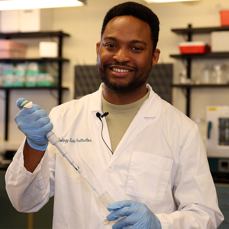 A researcher wearing a lab coat and gloves uses a pipette to transfer liquid into a sample tube while working in a laboratory.