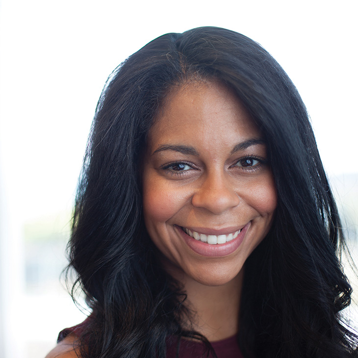 A person with long dark hair smiles toward the camera in a professional headshot taken against a softly blurred indoor background.