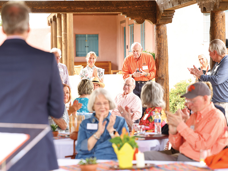 Attendees seated at tables under a covered courtyard applaud as a speaker addresses the group during an outdoor gathering at an adobe-style venue.