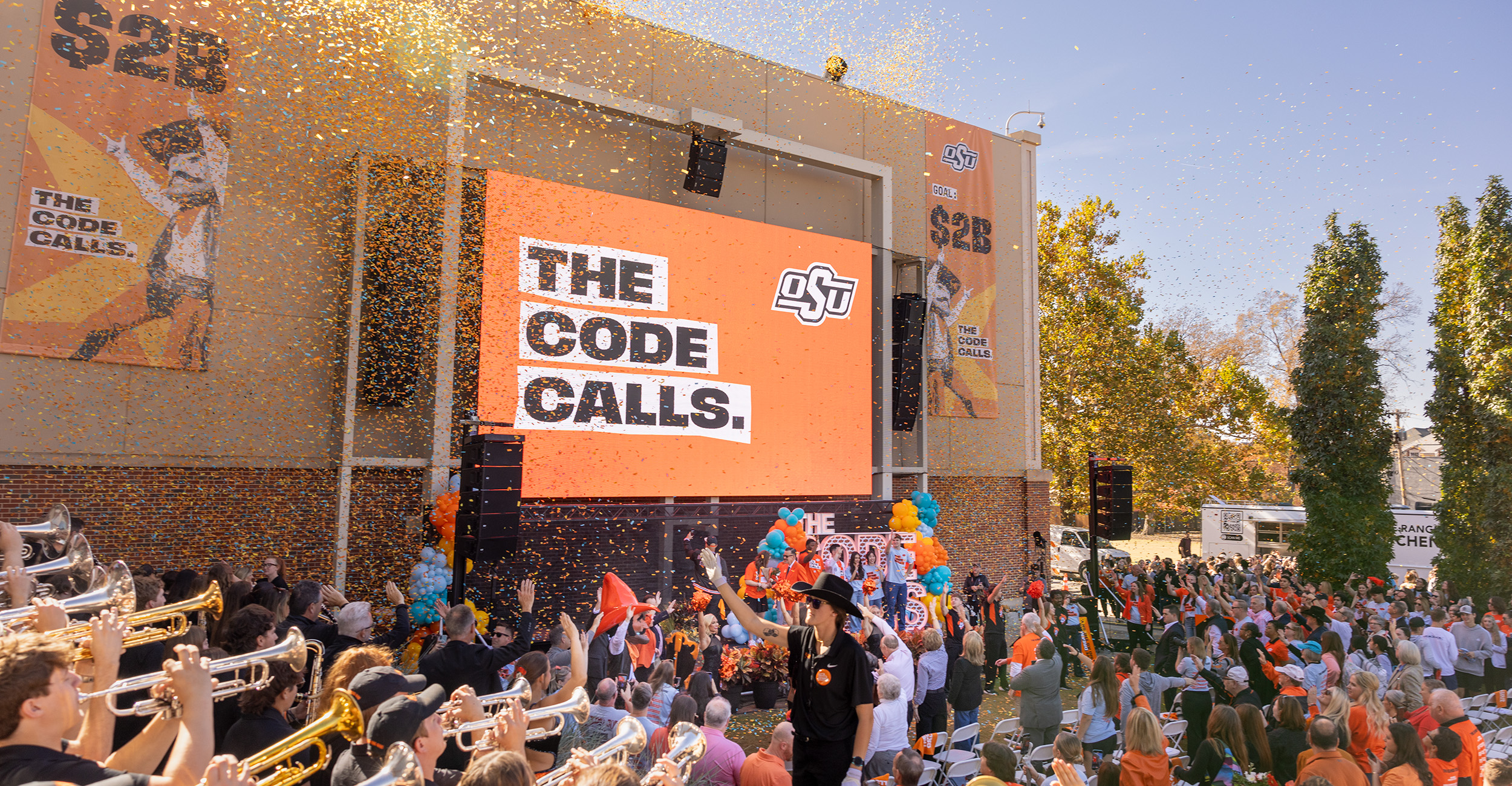 A large outdoor crowd gathers around a stage with a big screen displaying “THE CODE CALLS” and the OSU logo as confetti falls, with a marching band and speakers celebrating a campus campaign event.