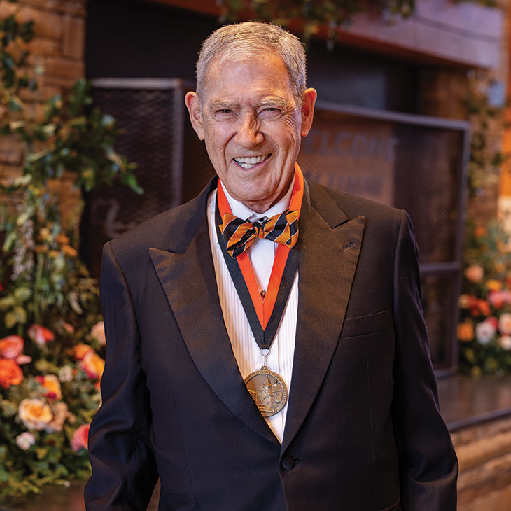 A formally dressed individual wearing a tuxedo, striped bow tie, and medallion stands in front of a stone fireplace and floral arrangements at an awards or recognition event.