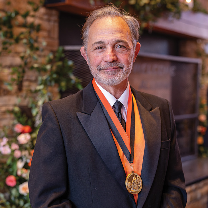 A formally dressed individual wearing a tuxedo and medallion poses in front of a stone fireplace and floral arrangements at an awards or recognition event.