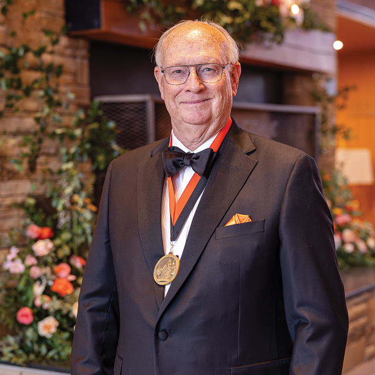 A formally dressed individual in a tuxedo with a bow tie and a medallion stands in front of a stone fireplace and floral arrangement at an awards or recognition event.