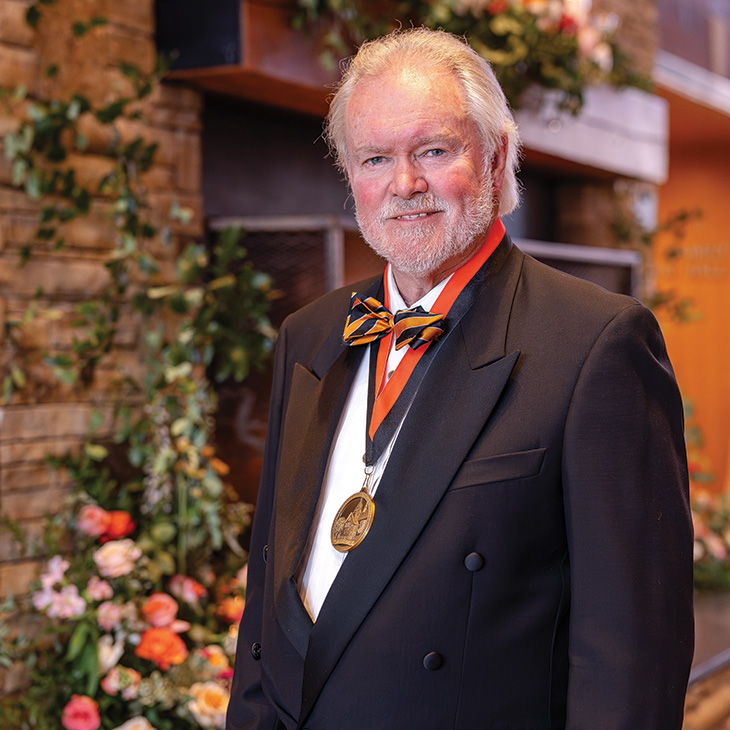 A formally dressed individual wearing a tuxedo, bow tie, and commemorative medallion stands in front of a stone fireplace and floral arrangements at an awards or recognition event.