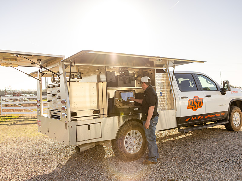 An Oklahoma State University veterinary field service truck with open side compartments is used by a person operating onboard diagnostic equipment in a rural setting.