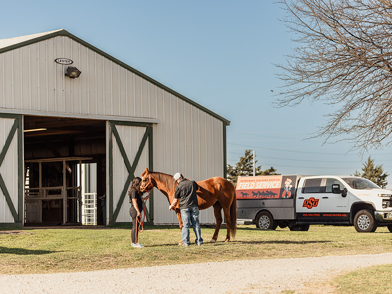 Two people stand beside a horse outside a barn while an Oklahoma State University veterinary field service truck is parked nearby, illustrating on-site equine care in a rural setting.