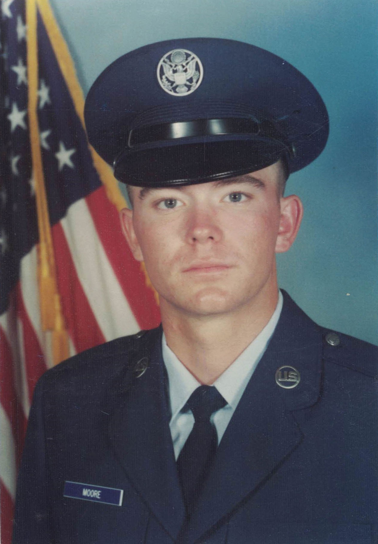 A young U.S. Air Force servicemember named Moore poses in dress uniform before an American flag in an official military portrait.