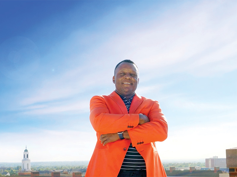 A person wearing a bright orange blazer and standing with arms crossed poses on a rooftop overlooking a city skyline under a clear blue sky.