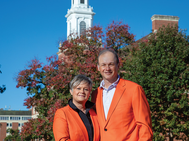 Two individuals wearing bright orange jackets stand together outdoors in front of a campus building and autumn-colored trees, posing for a portrait.