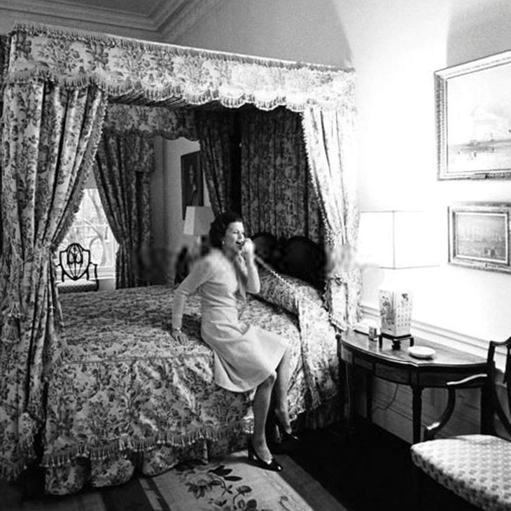 Black-and-white photograph of a person sitting on the edge of an ornate canopy bed in a decorated bedroom, holding a telephone near a bedside table with a lamp and framed pictures.