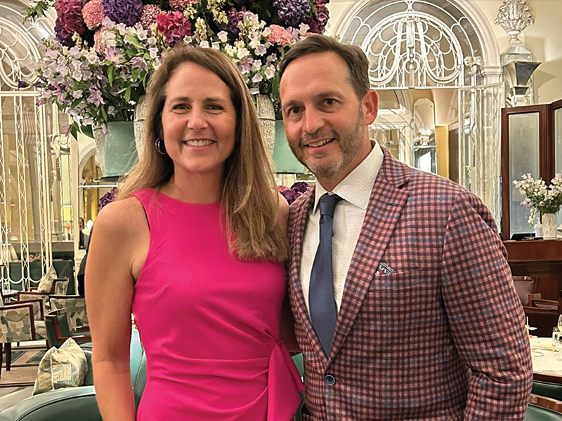 Two individuals dressed in formal attire stand closely together indoors in front of large floral arrangements, posing for a portrait at an event or reception.