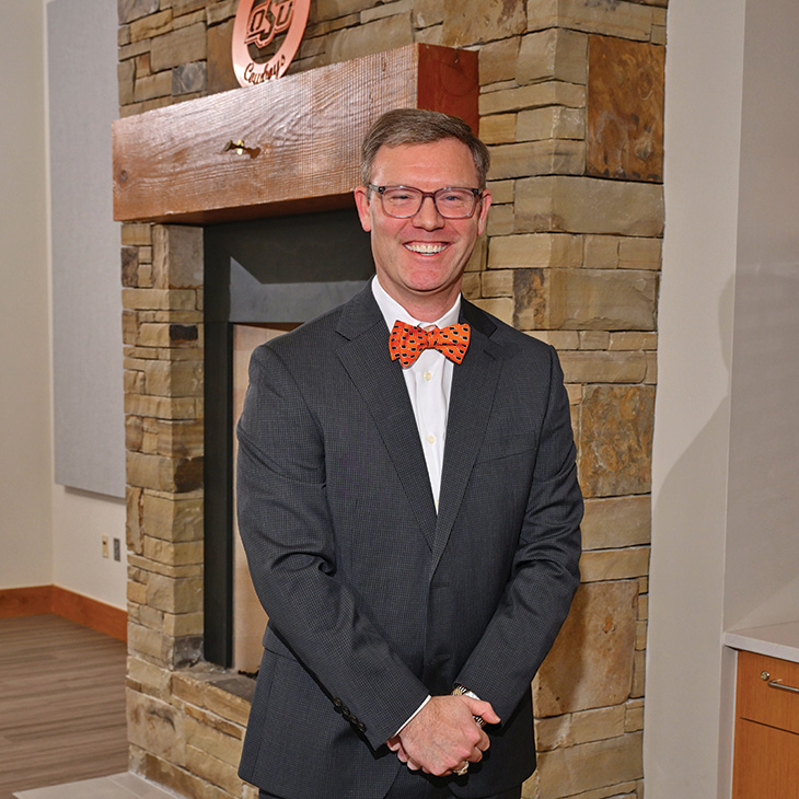 Color photograph of a person standing indoors in front of a stone fireplace, wearing a dark suit with an orange bow tie and glasses, facing the camera.