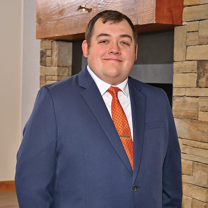 Color photograph of a person standing indoors in front of a stone fireplace, wearing a blue suit and orange tie, facing the camera.