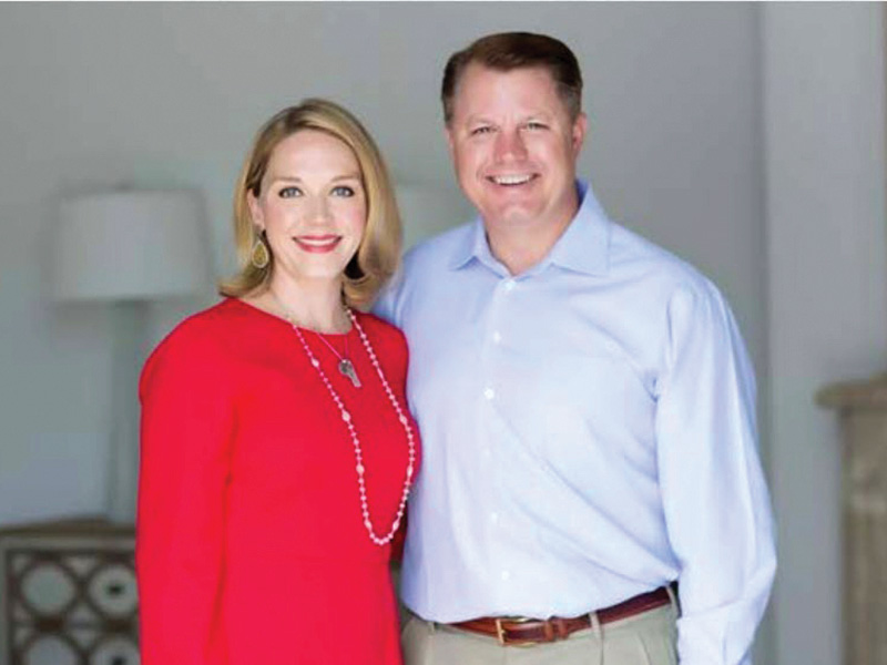 Two individuals stand side by side indoors, dressed in semi-formal attire and posing for a portrait against a softly lit neutral background.