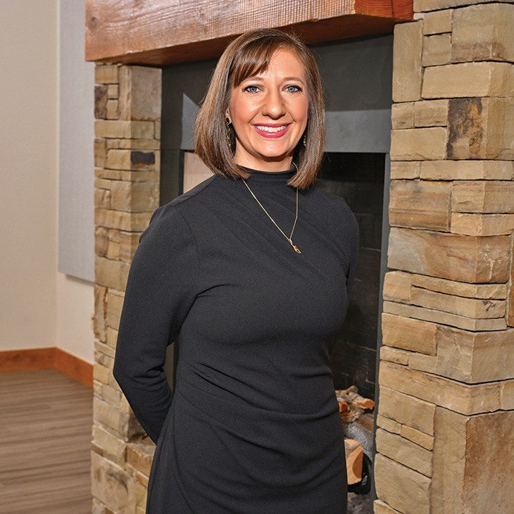 Color photograph of a person standing indoors in front of a stone fireplace, wearing a black dress and facing the camera.