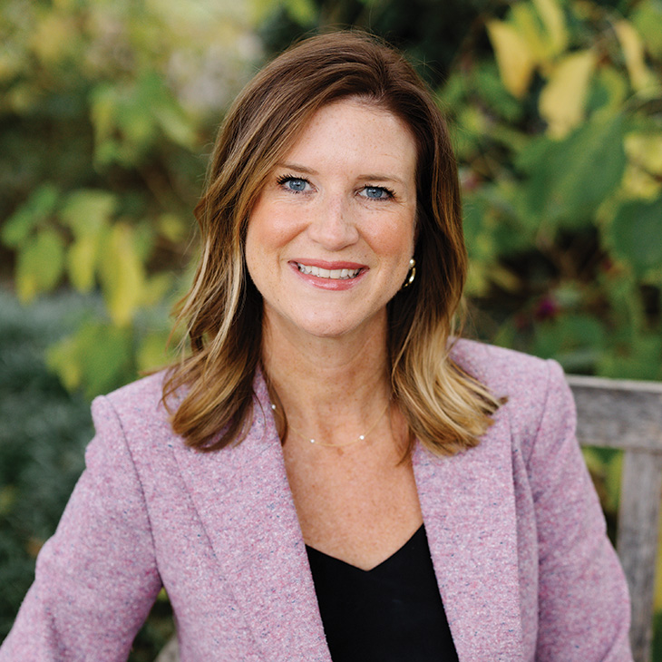 A person wearing a pink blazer sits outdoors on a bench in front of green foliage, posing for a professional portrait.
