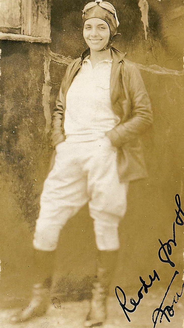 Sepia-toned historical photograph of an aviator wearing a leather helmet and goggles standing with hands in pockets against a rough wall.
