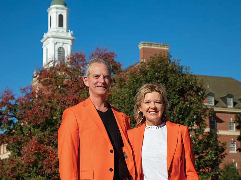 Two individuals wearing bright orange jackets stand outdoors in front of a campus building and autumn trees, posing for a portrait.