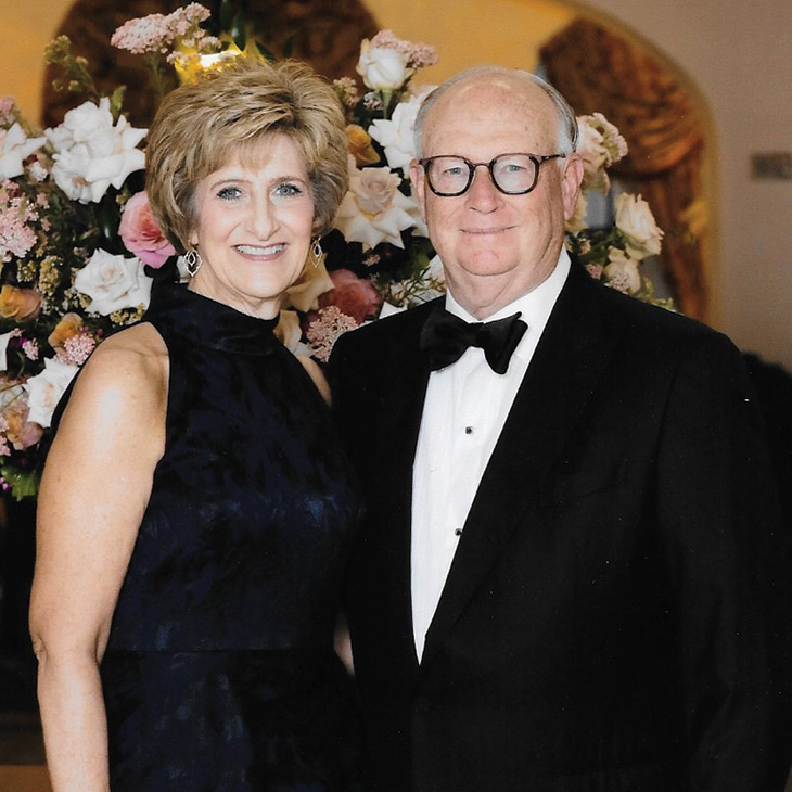 Two individuals in formal evening attire, including a tuxedo and a dark sleeveless dress, stand together in front of large floral arrangements, posing for a portrait at a formal event.