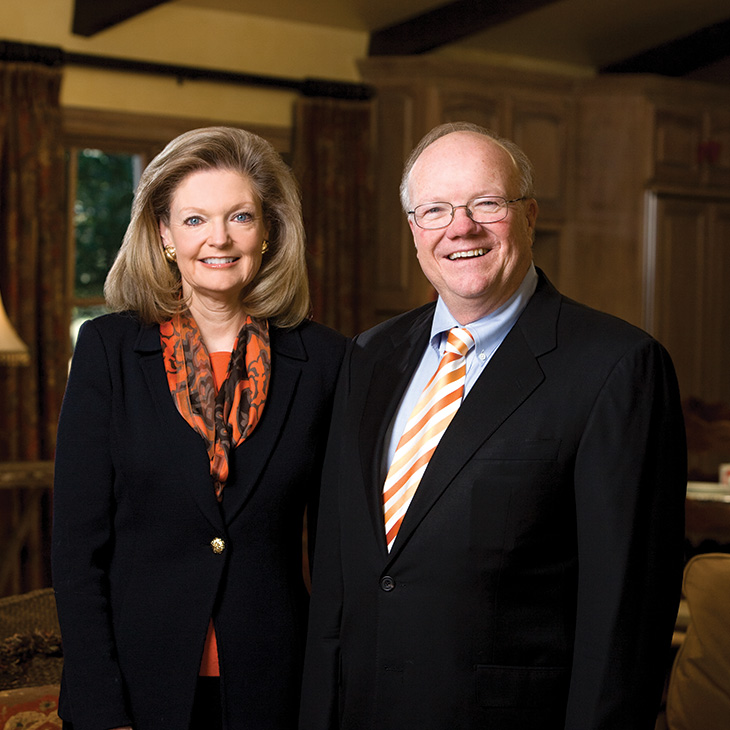 Two individuals in formal business attire stand side by side indoors, posing for a portrait in a warmly lit room with wood paneling and soft furnishings in the background.