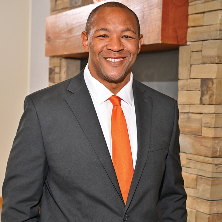 Color photograph of a person standing indoors in front of a stone fireplace, wearing a dark suit with a bright orange tie and facing the camera.
