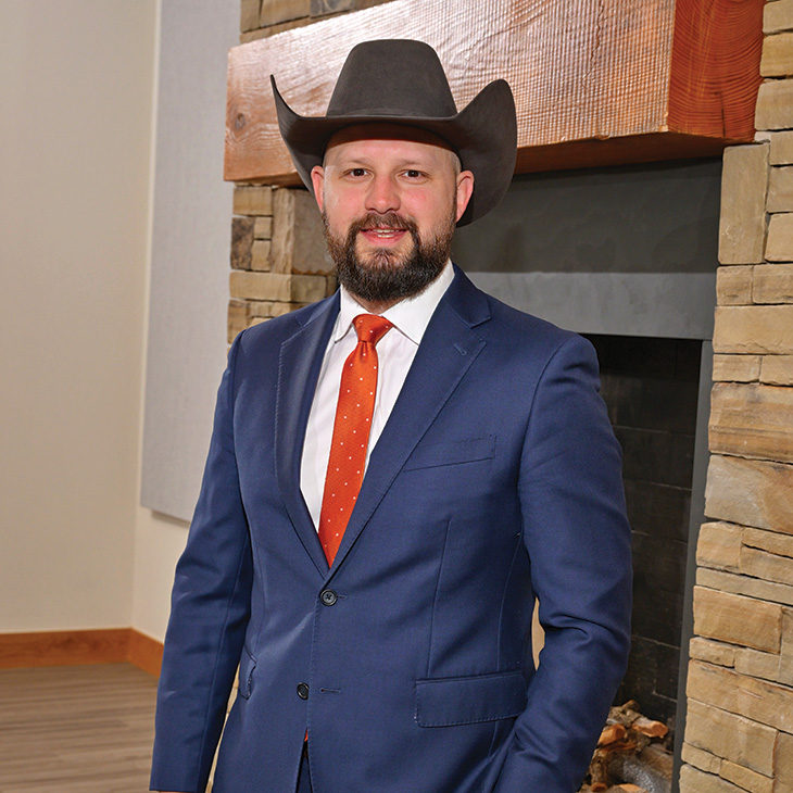 Color photograph of a person wearing a blue suit, white shirt, orange tie, and a cowboy hat standing indoors in front of a stone fireplace, facing the camera.