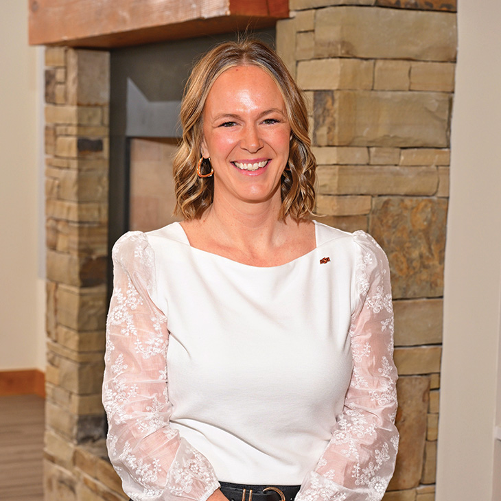 Color photograph of a person standing indoors in front of a stone fireplace, wearing a white blouse with lace sleeves and facing the camera.