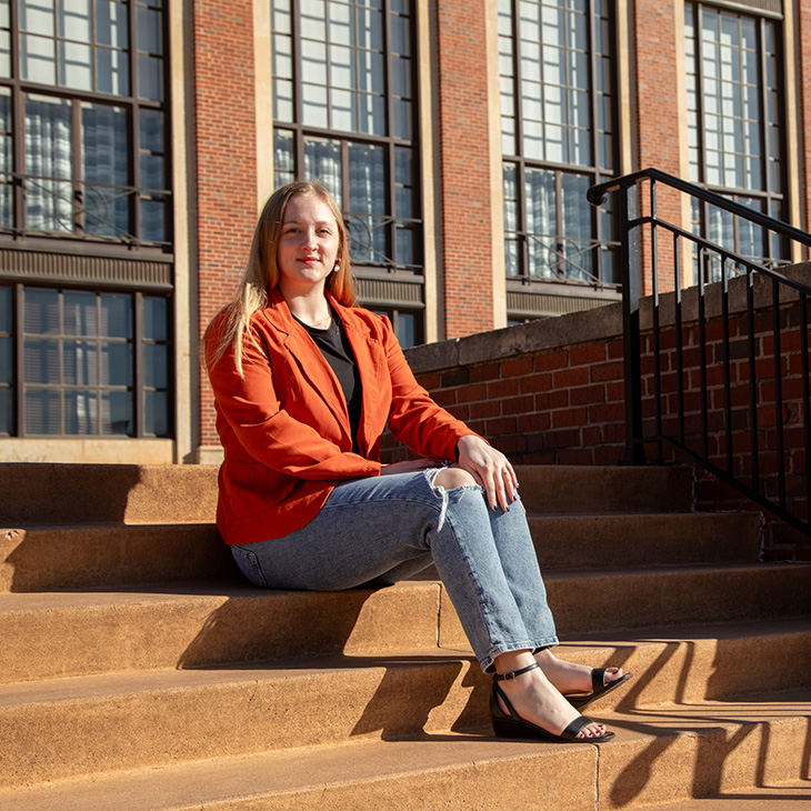 A person in an orange jacket sits on outdoor steps in front of a brick academic building, highlighting a campus setting.
