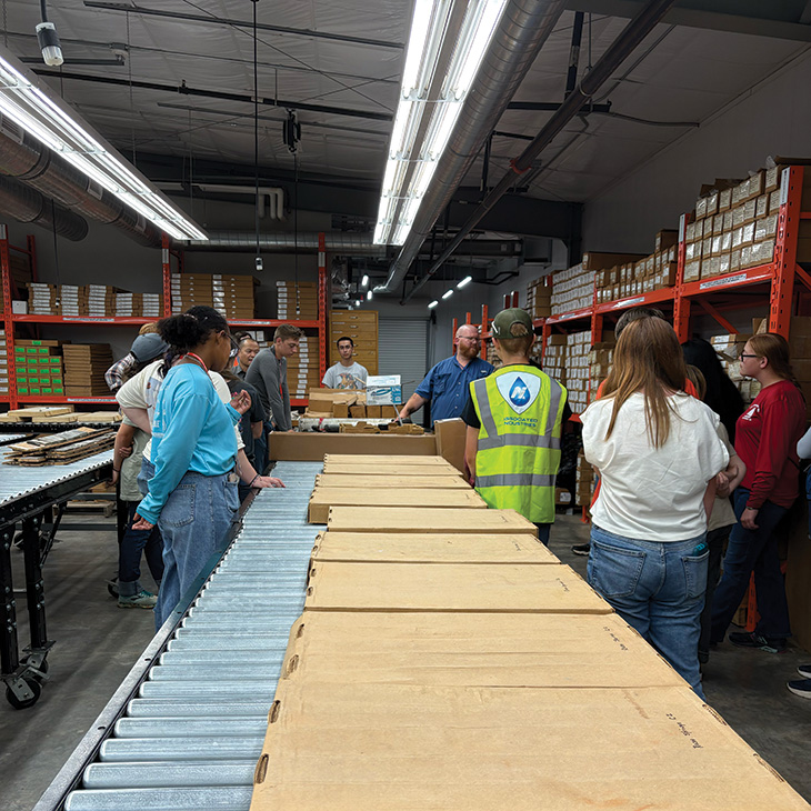 A group of people stands around a worktable in a warehouse lined with shelves of boxes, observing materials laid out on a conveyor system under bright overhead lights.