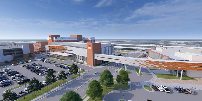 A wide aerial rendering shows a large, modern hospital complex with multiple interconnected buildings, elevated walkways, surrounding parking lots, and landscaped roadways.