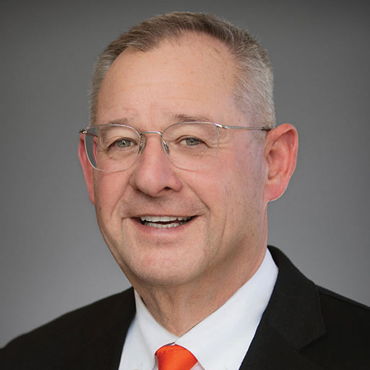 A person wearing glasses and a suit with a bright tie poses for a formal headshot against a neutral gray background.