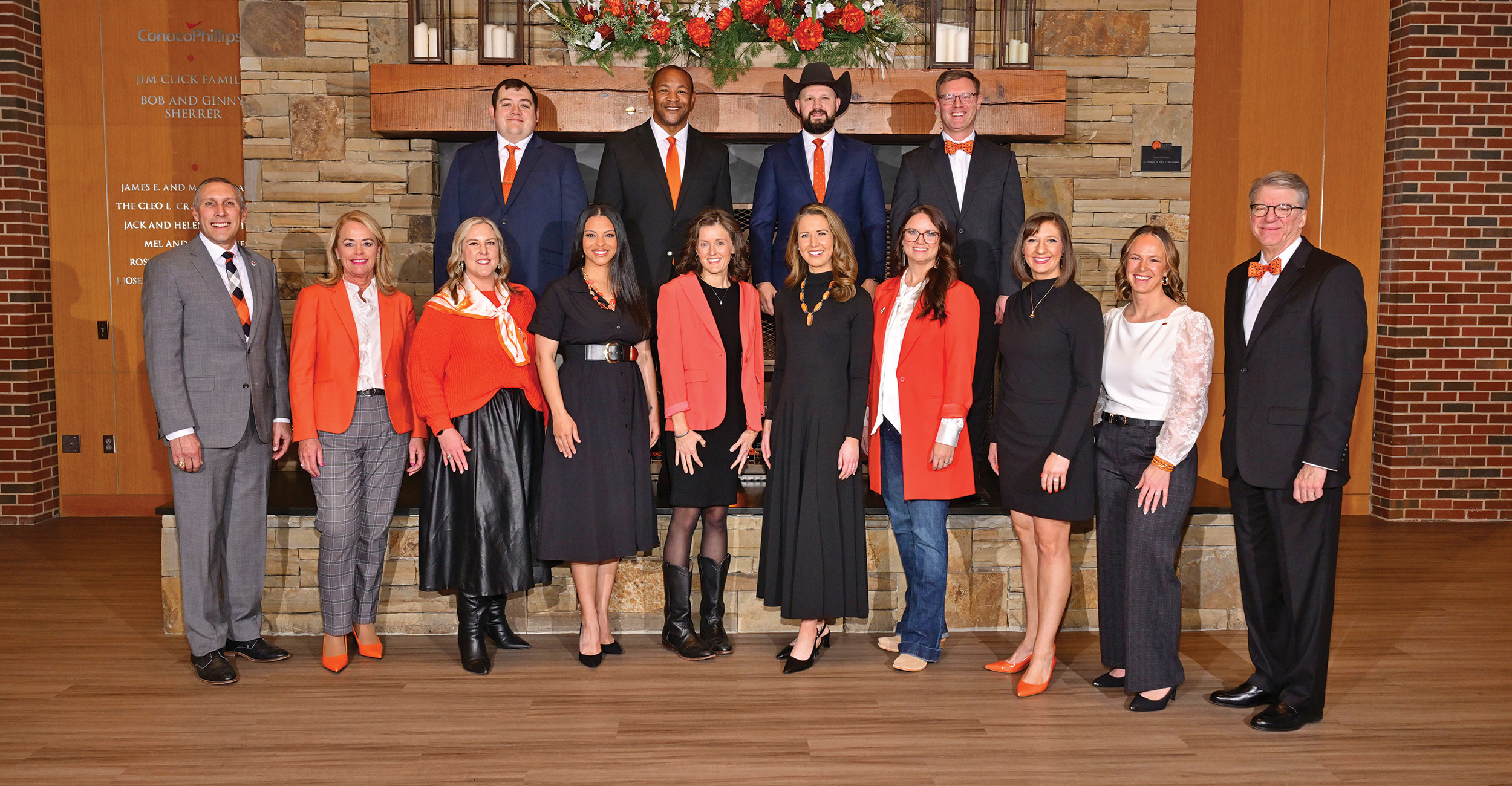 Color group portrait of multiple people dressed in formal attire, many wearing orange accents, standing in two rows in front of a stone fireplace with a decorative mantel.