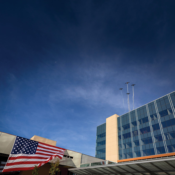A large American flag hangs beside a modern building as three small planes fly overhead in formation, leaving trails against a clear blue sky.