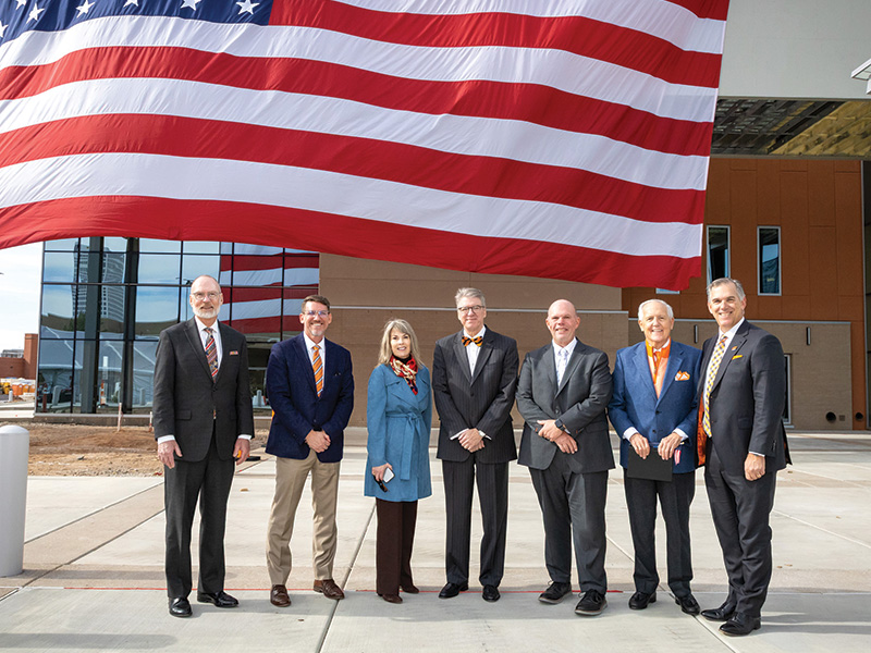 Six individuals stand in a row outdoors in front of a large suspended American flag at a modern building, posing for a formal group photo.