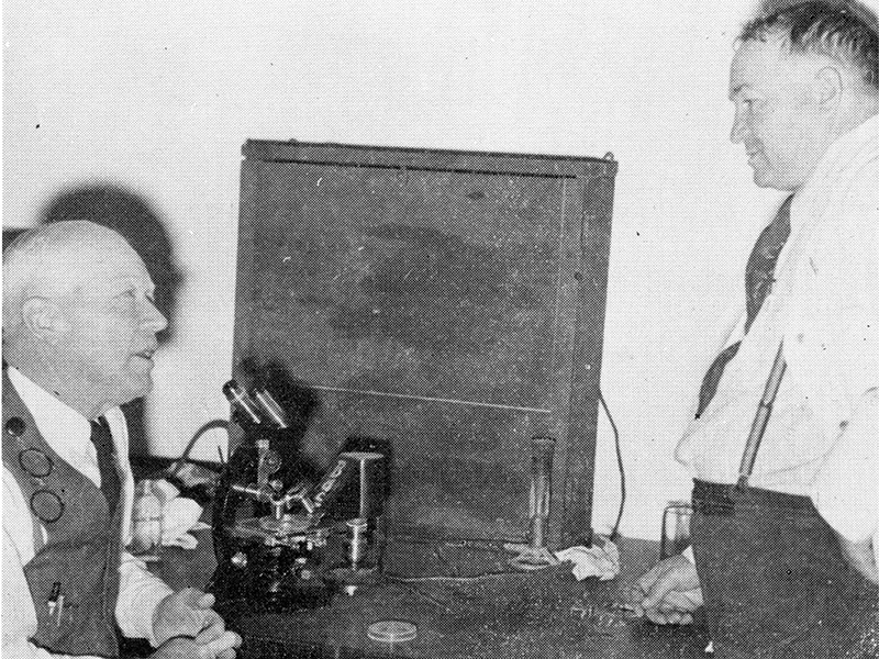 Two people in a black-and-white photo, seated and standing near a table with a microscope and scientific equipment.
