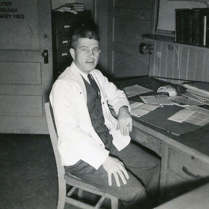 Person wearing a white lab coat seated at a wooden desk with papers and books in what appears to be an office or laboratory setting.