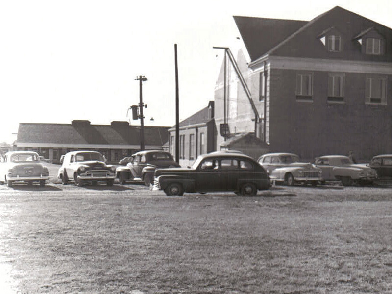 Black-and-white photo of several vintage cars parked near a large brick building and smaller structures.