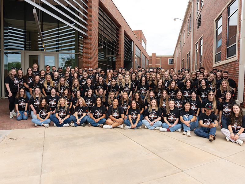 A large group of people wearing matching black T-shirts with a graphic design, posing for a group photo outside a modern brick building. They are arranged in several rows, some seated on the ground and others standing behind.