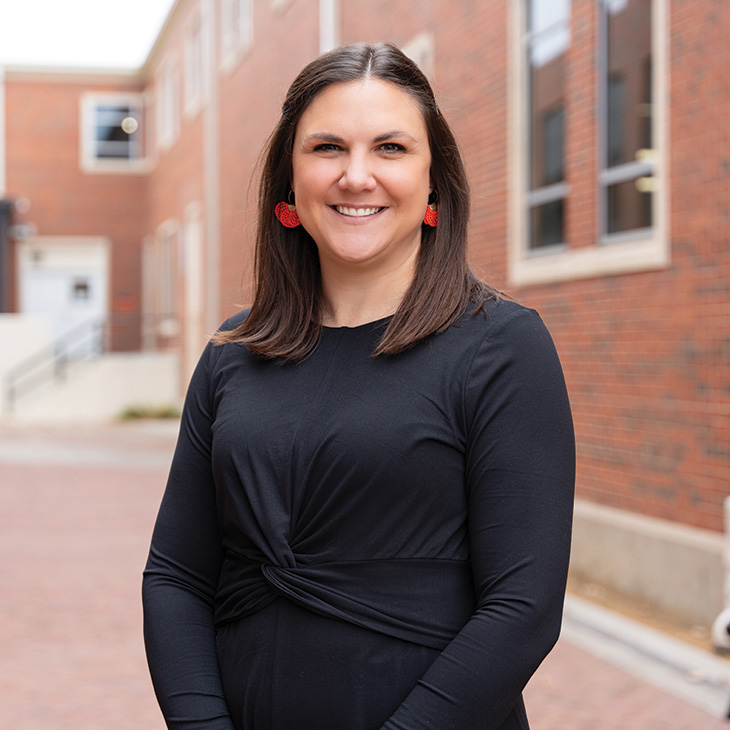 A person standing outdoors in a brick-paved walkway between red brick buildings. They are wearing a long-sleeve black dress with a knotted detail at the waist and red statement earrings. Their hands are clasped in front.