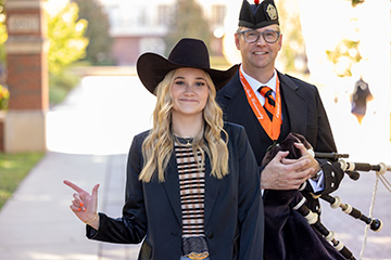 A man wearing a formal Scottish outfit with an Oklahoma State University medal walks beside a woman in a black cowboy hat and blazer. They are outdoors on a sunny day with trees and other people in the background.