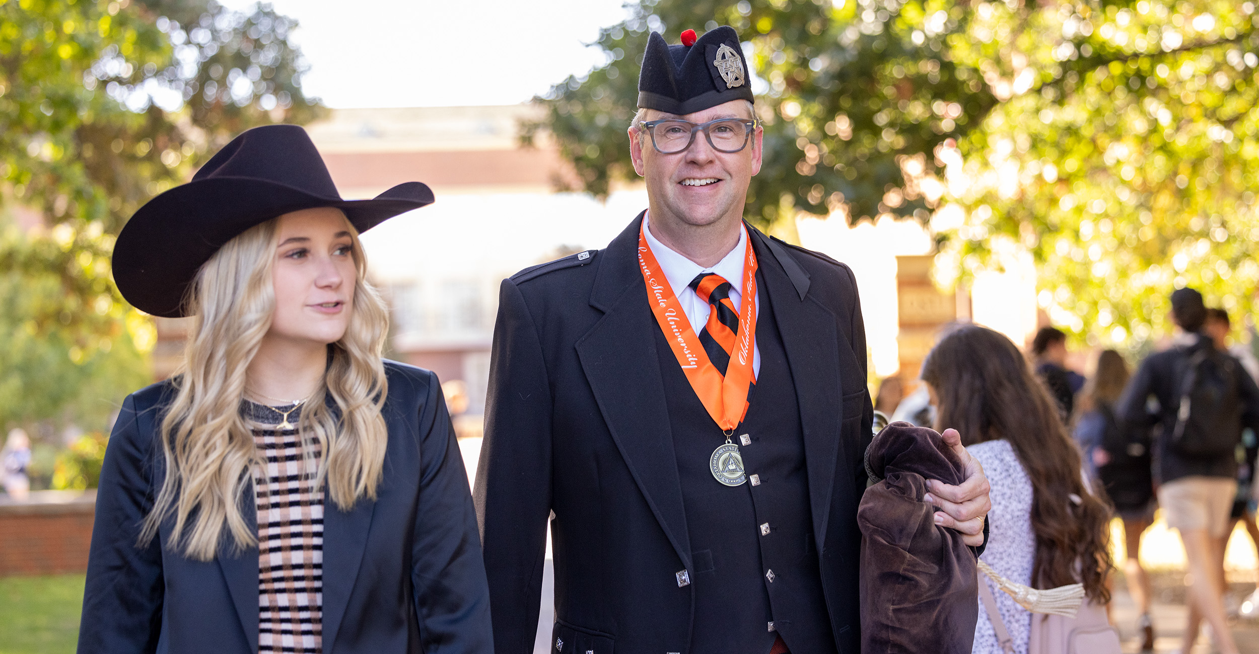 A man wearing a formal Scottish outfit with an Oklahoma State University medal walks beside a woman in a black cowboy hat and blazer. They are outdoors on a sunny day with trees and other people in the background.